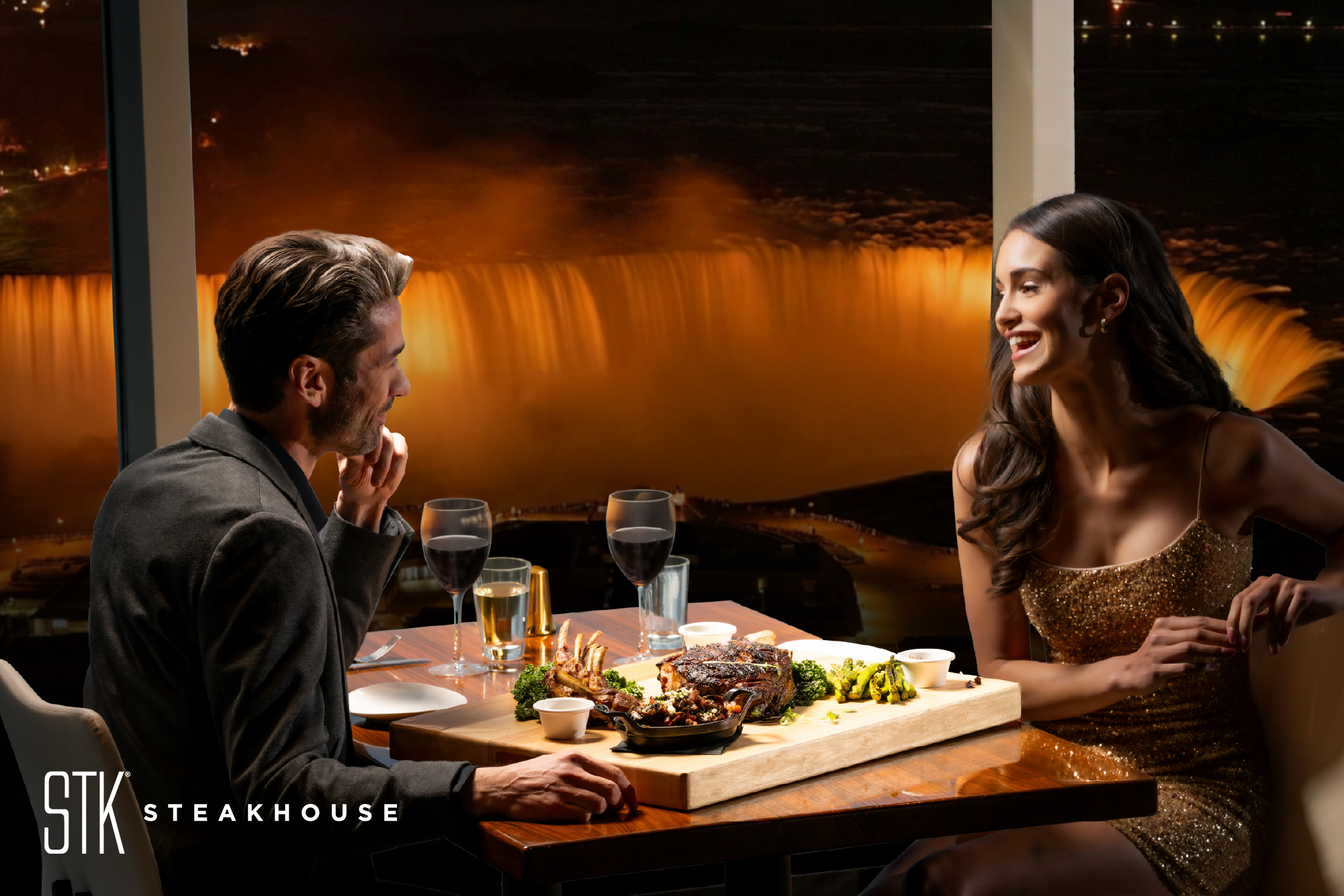 Couple happily conversing at table in STK Steakhouse Falls View seated directly beside the window viewing Niagara Falls with wine, steak and seafood on the table.